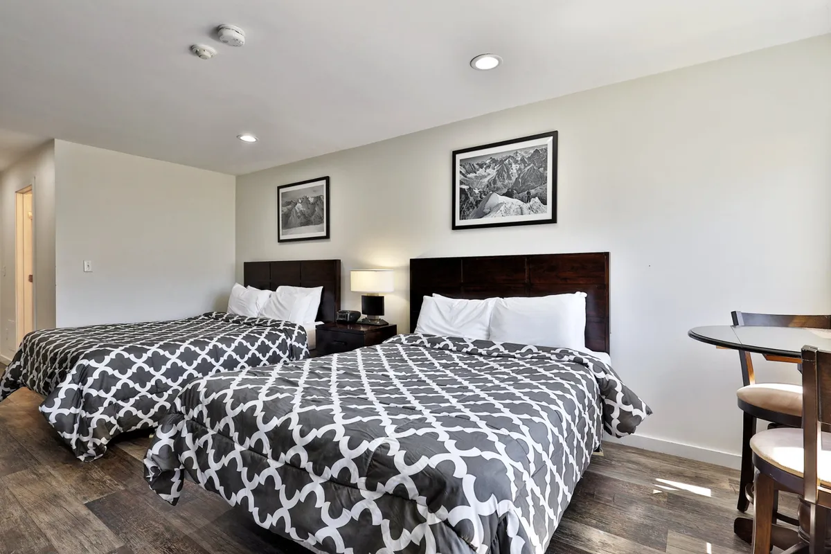 The image showcases a bright, inviting studio interior with two plush queen beds, stylish wall art, and a cozy dining nook—perfect for morning coffee by the window. The overall atmosphere is warm and comfortable, ideal for relaxing or connecting after a day on the slopes.