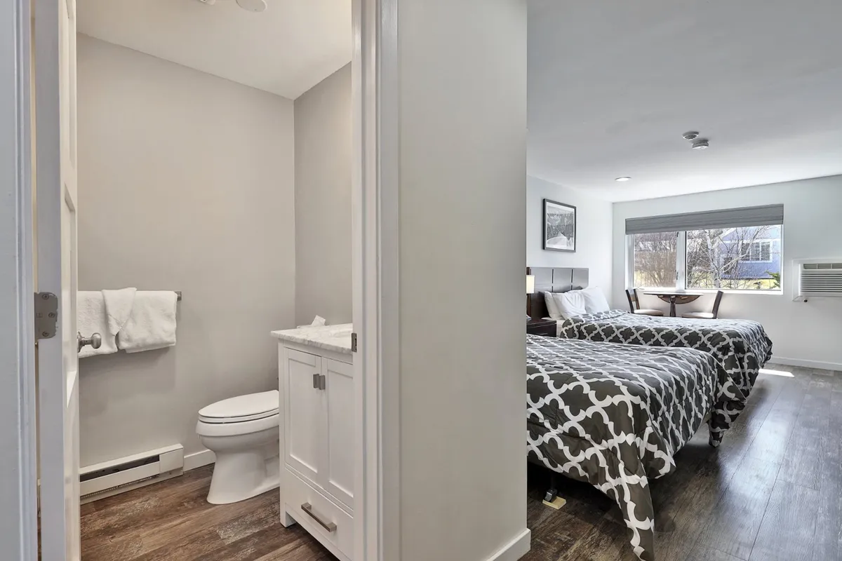 A bright studio living space with two beds adorned in patterned bedding, a bathroom entrance, and a window streaming in natural light. The room feels spacious and comfortable, ideal for relaxing after exploring.