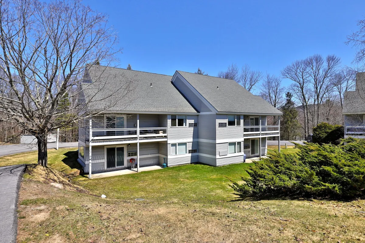 The exterior shows a multi-unit building with a wide green lawn and mature trees, evoking a peaceful suburban setting. The lawn is suitable for outdoor gatherings, picnics, or casual strolls—perfect for guests planning to spend time outside during the summer months.