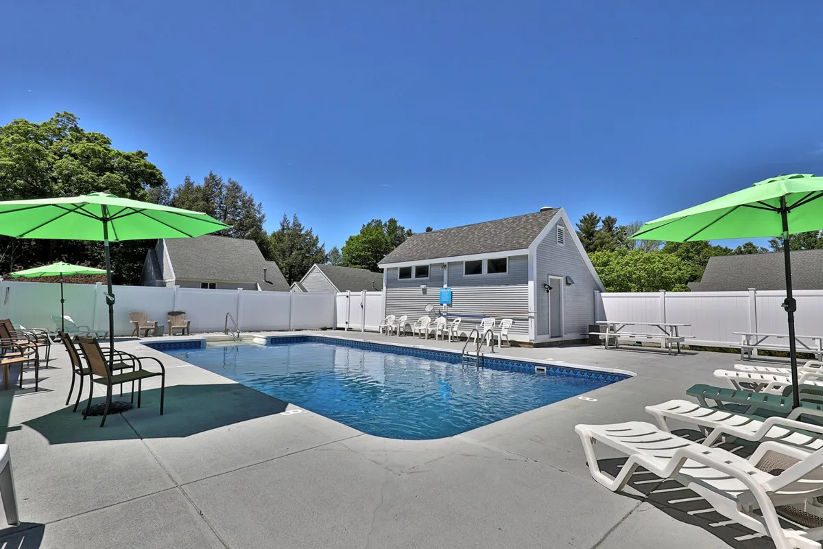 This lively poolside scene features the bright blue swimming pool, green umbrellas, lounge chairs, and is framed by lush trees. The changing room building is visible, underscoring convenience for poolside guests. The mood is relaxed and cheerful, ideal for summer enjoyment.