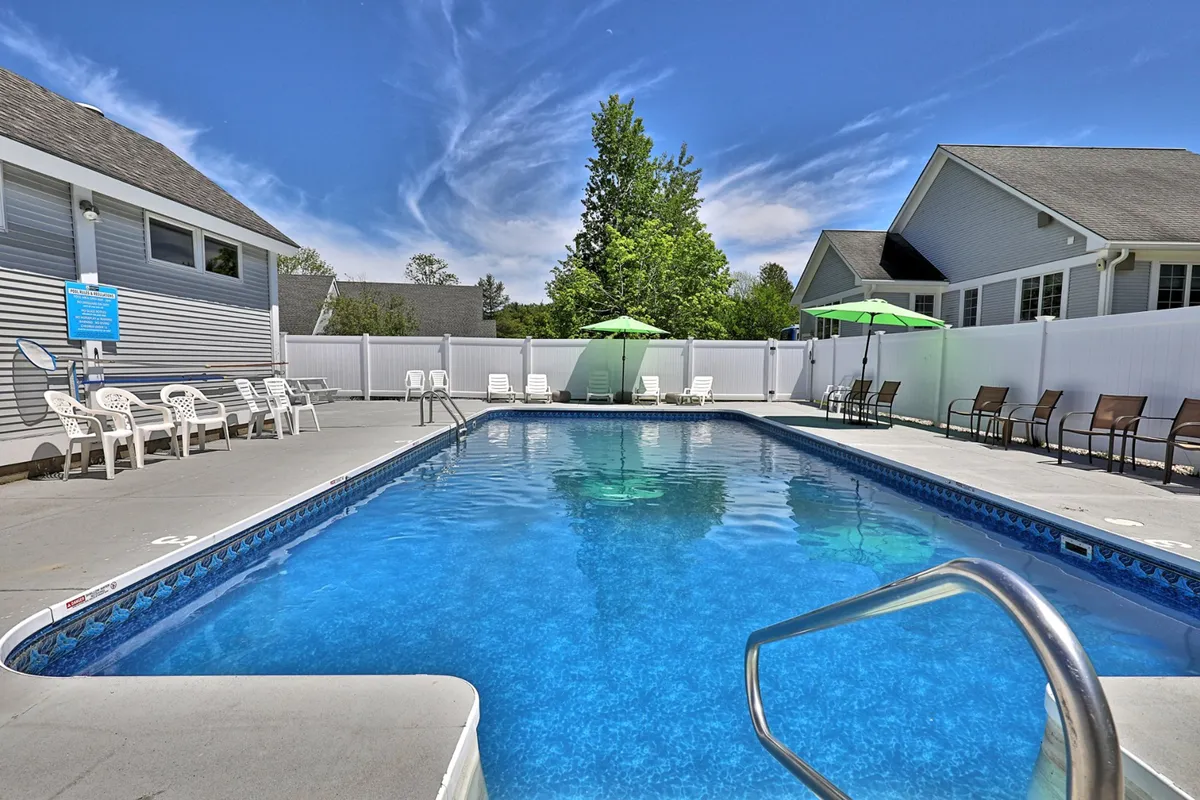 This vibrant exterior image captures the heated saltwater swimming pool surrounded by lounge chairs, green umbrellas, and lush trees. Sunlight glints on the water and people are seen swimming, sunbathing, and socializing, embodying the lively yet relaxing ambiance of summer at the complex.