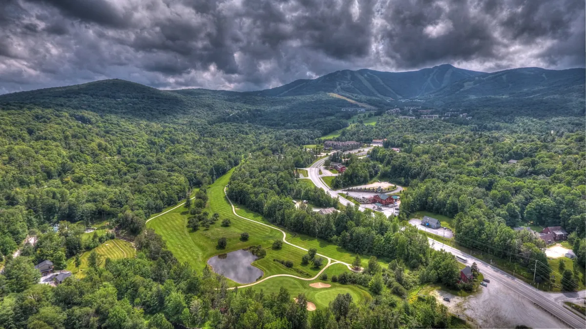 A panoramic vista featuring a majestic mountain range, a winding road, a golf course, and a tranquil pond surrounded by dense forest. The view is expansive and adventurous.